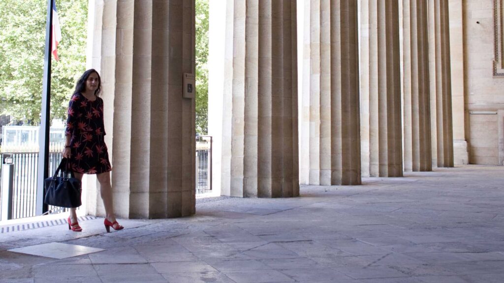 Mode Parisienne : Femme marchant sous une colonnade Femme en robe noire et rouge marchant sous une colonnade classique, portant un sac à main et des talons rouges.